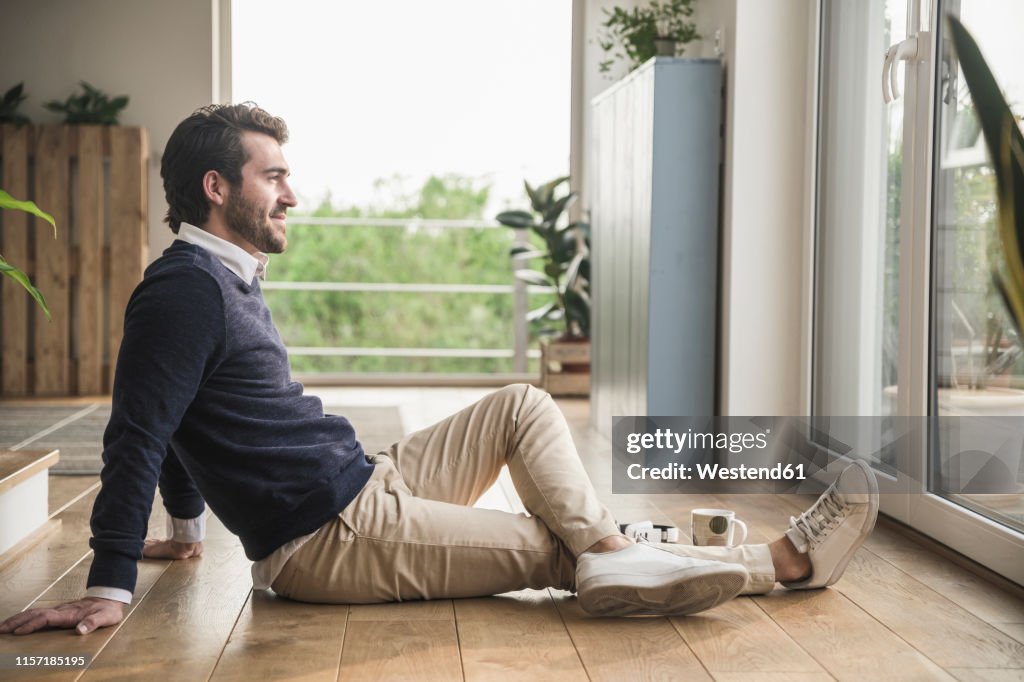Young man sitting on floor, looking out of window, relaxing