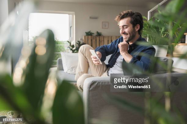 young man sitting on couch at home, using smartphone - sofa stock-fotos und bilder