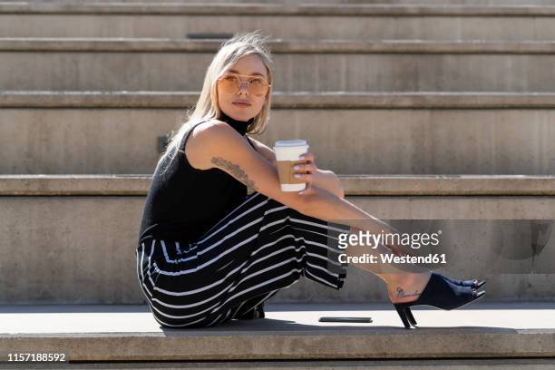 bortrait of blond young woman with coffee to go sitting on stairs outdoors - broekrok stockfoto's en -beelden