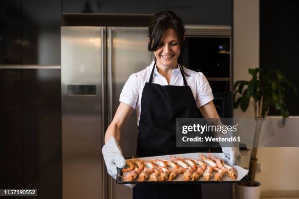 smiling woman holding a baking tray with shrimps in kitchen - seafood platter stockfoto's en -beelden