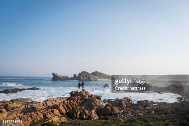 paar holding handen staan op rotsachtige kustlijn op het strand tegen sky - middlebare afstand stockfoto's en -beelden