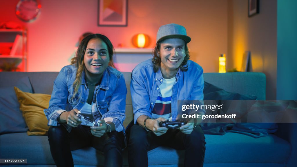Aufgeregt Black Gamer Girl und Young Man Sitzen auf einer Couch und Spielen VonVideo-Spiele auf Der Konsole. Sie spielen mit Wireless Controllern. Das Cozy Room ist mit warmem und Neonlicht im Licht.