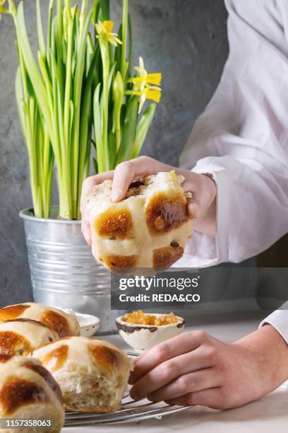Child hands take homemade Easter traditional hot cross buns on cooling rack on white marble table with narcissus flowers.