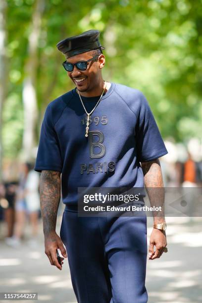 Basket-ball player Carmelo Anthony wears a leather hat, a blue "1895 B Paris" top, blue pants, a necklace, reflective sunglasses, a golden watch,...