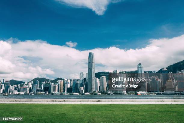 panoramic view of the iconic hong kong city skyline by the promenade of victoria harbour against clear blue sky and meadow - hong kong central stock pictures, royalty-free photos & images