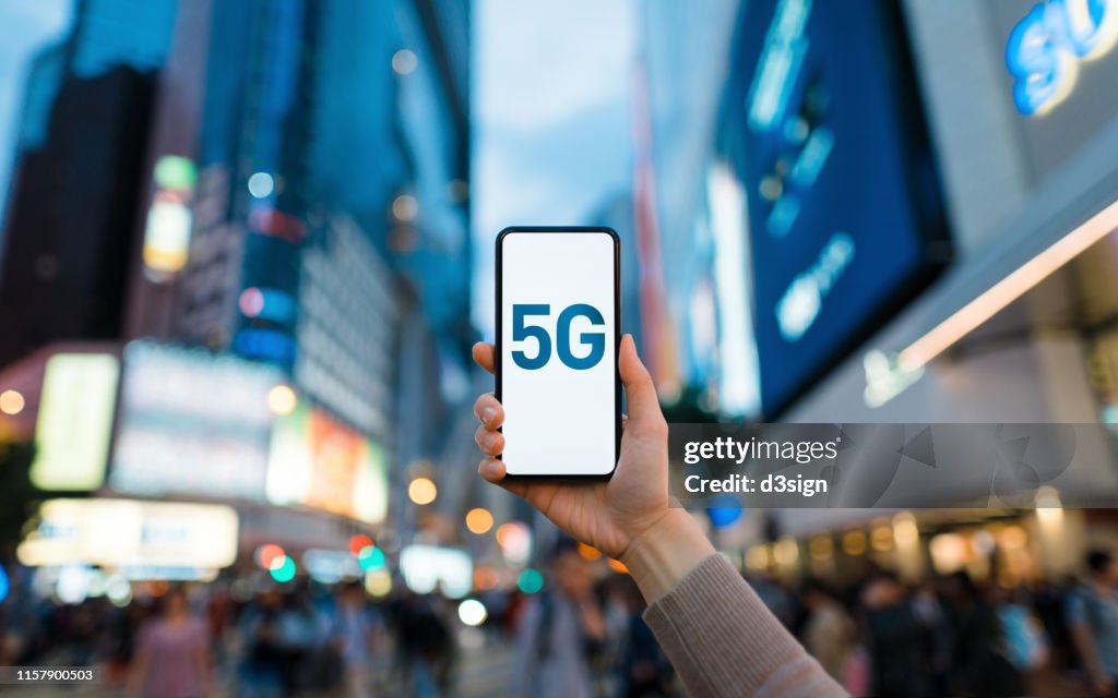 Woman's hand using smartphone against illuminated financial district in the city, with the concept of 5G communications technology