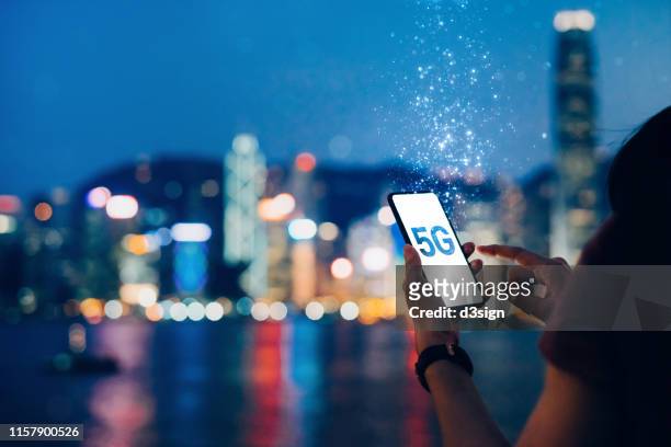 young woman using smartphone against the iconic city skyline of hong kong by the promenade of victoria harbour at night, with the concept of 5g communications dissolved into light particles - 通信設備 ストックフォトと画像