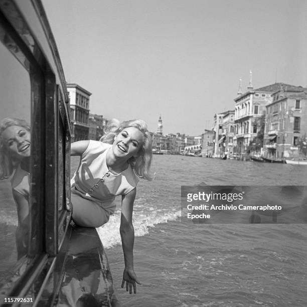 American actress Carroll Baker, wearing a white dress, portrayed while leaning out of a water taxi on the Canal Grande, Rialto Bridge in the...