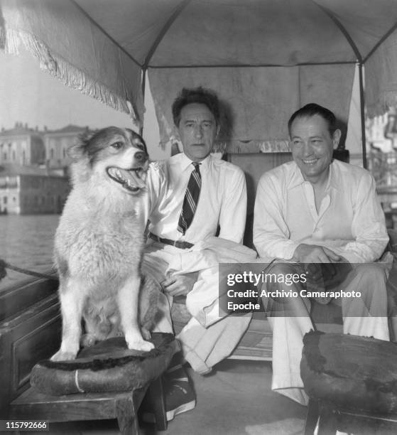 French writer Jean Cocteau with a striped tie, sitting on a covered gondola with Roberto Rossellini and a dog, floating on the Canal Grande, Venice...