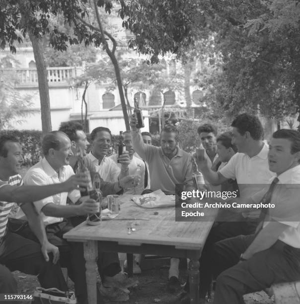 American actor Paul Neuman, sitting at a table in the Excelsior garden with many other men, holding a bottle of beer and toasting with them, Lido,...