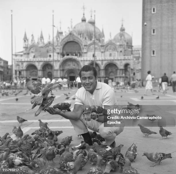 Japanese actor Toshiro Mifune smiling, crouched in St. Mark Square, feeding pigeons and posing for the picture, Venice, September 1961.
