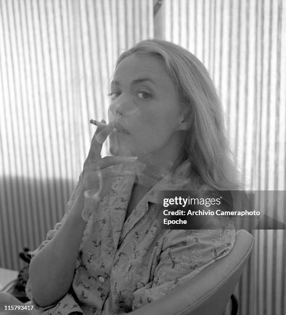 French actress Jeanne Moreau, wearing an embroidered shirt, portrayed while smoking a cigarette and making smoke spirals, a striped curtain in the...