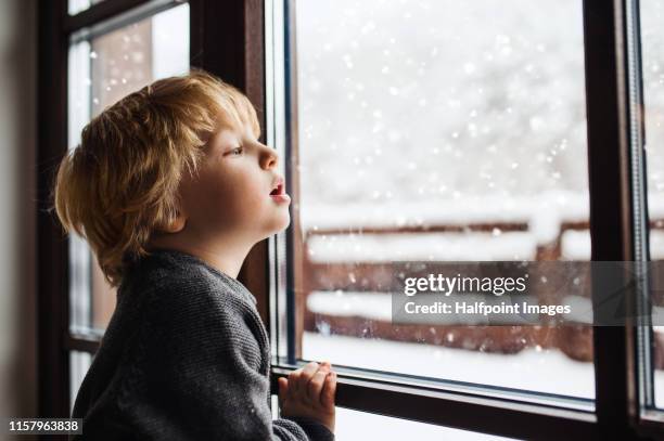 a small bored boy indoors standing by the window, looking out. - snow window stock pictures, royalty-free photos & images