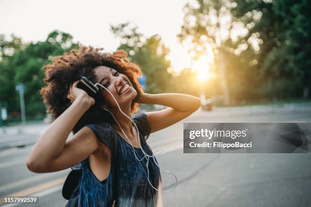 junge erwachsene frau mit afro-haar tanzen in der stadt bei sonnenuntergang beim hören von musik - smartphone musik stock-fotos und bilder