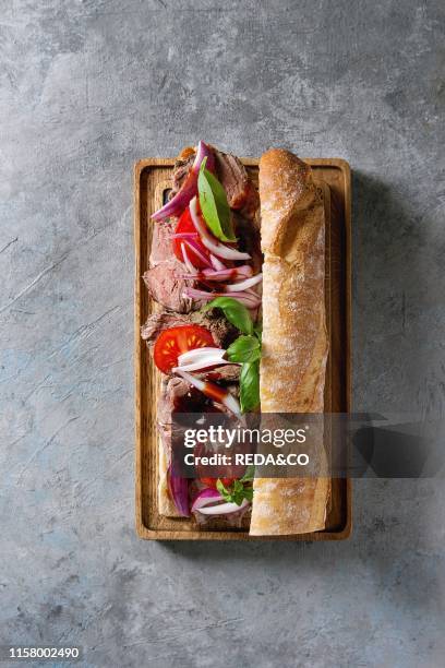 Beef baguette sandwich with tomatoes. Basil. Red onion served on wooden serving board over grey texture background. Flat lay. Space.