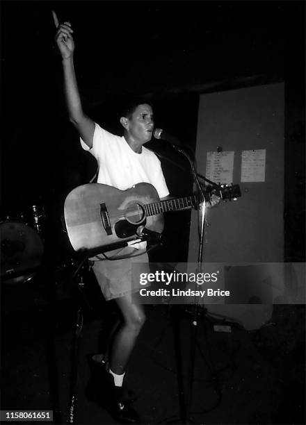 Susan Gottlieb, known as Phranc, on acoustic guitar, raises right hand pointing upward while standing and singing into mic at Free 2 Fight event of...