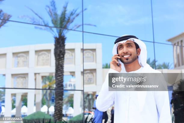 a handsome arab model speaking in phone happy smiling and wearing uae traditional dress - bahrain man stock pictures, royalty-free photos & images