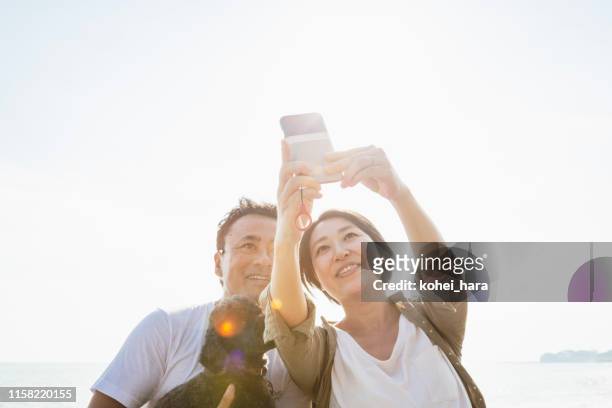 aziatisch stel met hond die selfie in het strand neemt - mid adult couple stockfoto's en -beelden