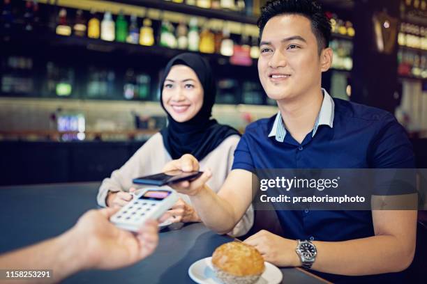 young malaysian couple is paying using mobile phone in the cafe - islamic finance stock pictures, royalty-free photos & images