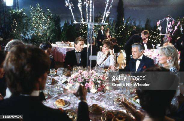 Stars on television hit show "Dynasty" sit at table during filming of banquet scene in studio in Hollywood circa 1985: