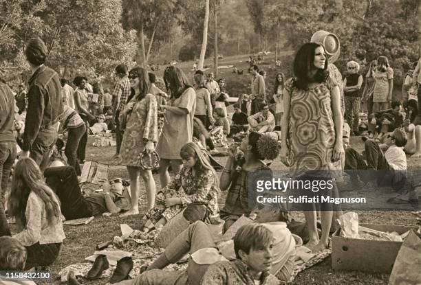 Three women in "mod" shifts standing, man reading Proust lying on the grass, many people relaxing on the grass at the 1st Elysian Park Love-In on...