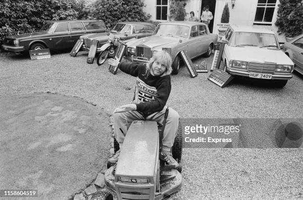 English keyboardist, songwriter, producer, television and radio presenter, and author Rick Wakeman on a compact tractor in the front yard of a house,...