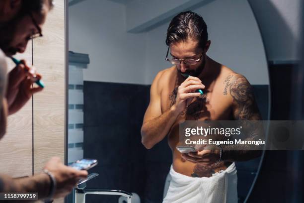 mid adult man with tattoos brushing teeth and looking at smartphone at bathroom mirror, mirror image - brush teeth phone stock pictures, royalty-free photos & images