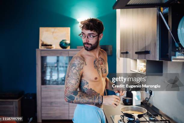 mid adult man with tattooed chest preparing breakfast while looking over shoulder - distrarsi foto e immagini stock