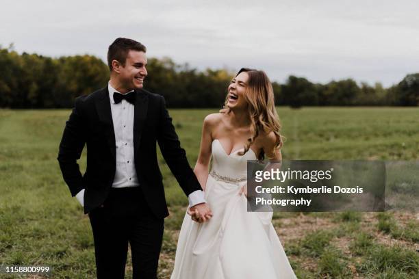 romantic young bride and groom holding hands and laughing in field - newly married stock pictures, royalty-free photos & images