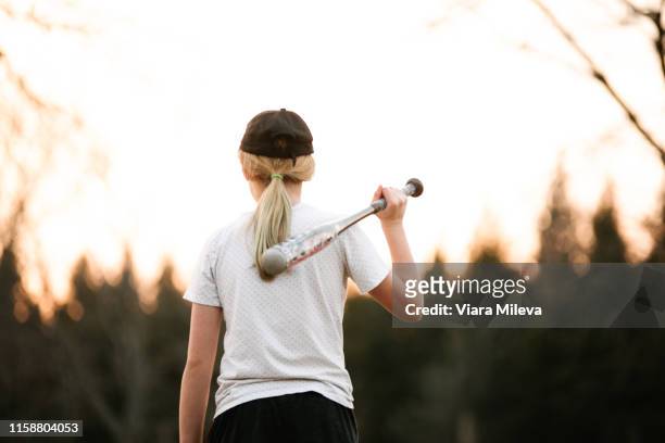 girl with baseball bat over her shoulder in rural field, rear view - girl baseball cap stock pictures, royalty-free photos & images