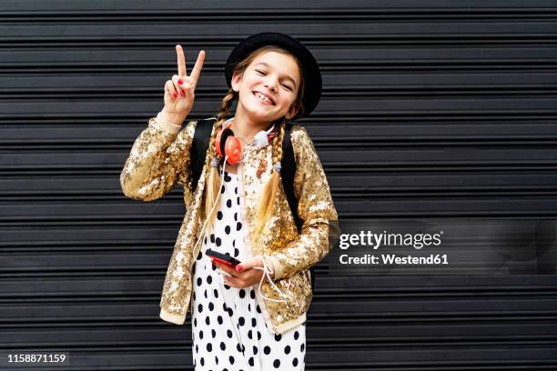 portrait of happy girl with smartphone wearing hat and golden sequin jacket showing victory sign - victory sign stock-fotos und bilder