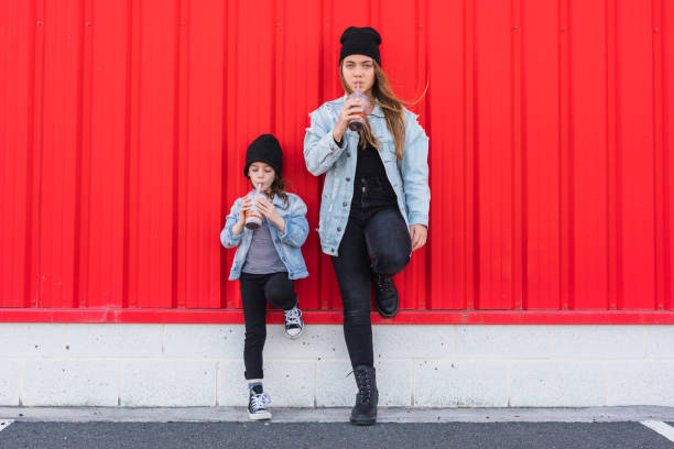 teenage girl and little sister leaning against red wall drinking cocoa - teens sizes stock pictures, royalty-free photos & images