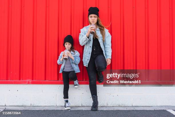 teenage girl and little sister leaning against red wall drinking cocoa - matching outfits stock pictures, royalty-free photos & images