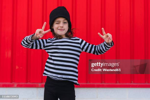 portrait of winking little girl wearing striped shirt and black cap showing victory sign - black shirt 個照片及圖片檔