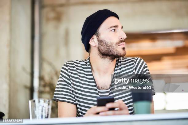 young man with cell phone wearing a beanie looking around - coffee chat stockfoto's en -beelden