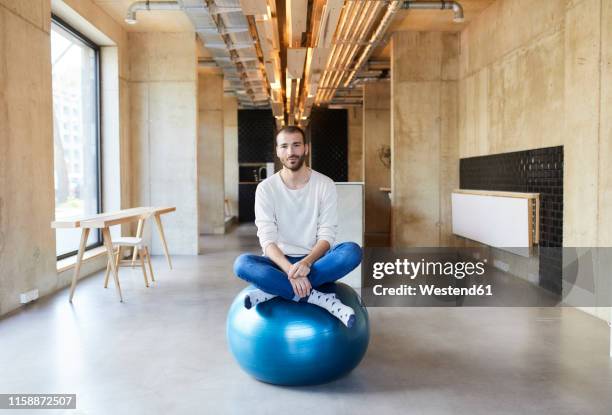 portrait of young man sitting on fitness ball in modern office - yoga ball work stock pictures, royalty-free photos & images