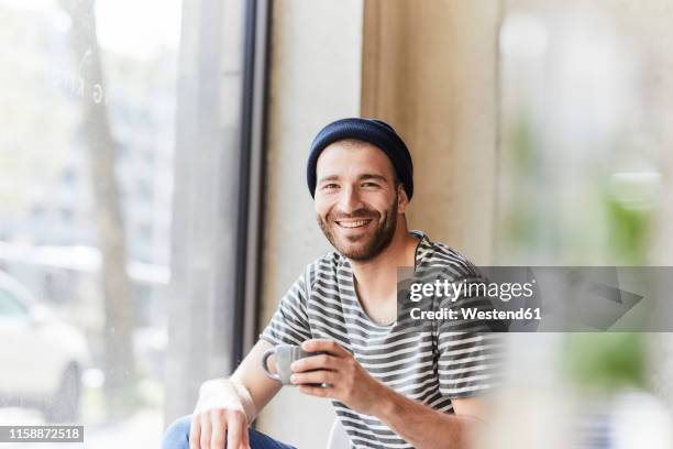 portrait of smiling young man holding coffee cup at the window - mann lachen blick in die kamera stock-fotos und bilder