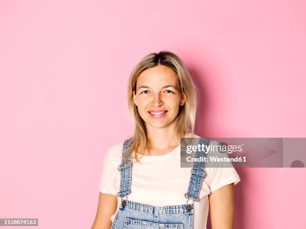 portrait of smiling blond woman wearing dungarees in front of pink background - jeans latzhose frau stock-fotos und bilder