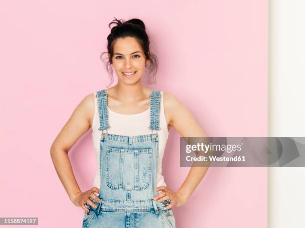 portrait of young smiling woman with black hair and hands on hips in front of pink background - jeans latzhose frau stock-fotos und bilder
