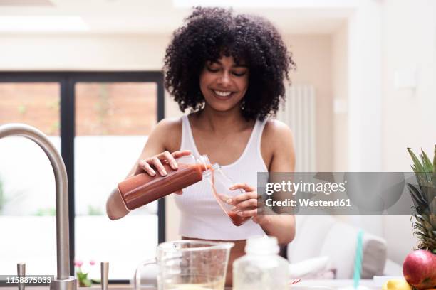 smiling young woman preparing healthy smoothie in kitchen - bottles glass top stock pictures, royalty-free photos & images