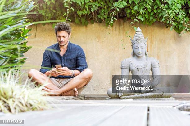 man sitting cross-legged next to buddha statue in a zen garden, using smartphone - zen garden stock pictures, royalty-free photos & images