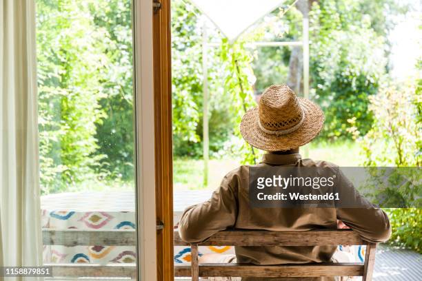 back view of senior man wearing straw hat on terrace enjoying his garden - strohoed stockfoto's en -beelden