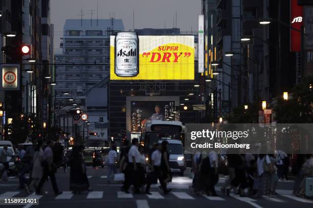 Pedestrians walk across a road as a billboard for Asahi Breweries Ltd.'s Asahi Super Dry beer stands illuminated at dusk in Tokyo, Japan, on...