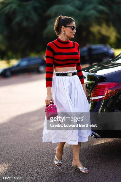 Laetitia Casta wears large earrings, sunglasses, a red and black cropped bare belly top, a belt, a white skirt with embroidery, a pink bag, silver...