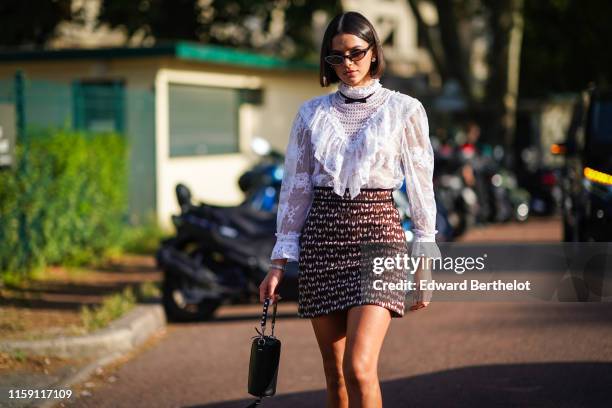 Bruna Marquezine wears earrings, a white lace embroidered top, a skirt, a balck bag with fringes, black boots, a small bow tie, outside Miu Miu Club...