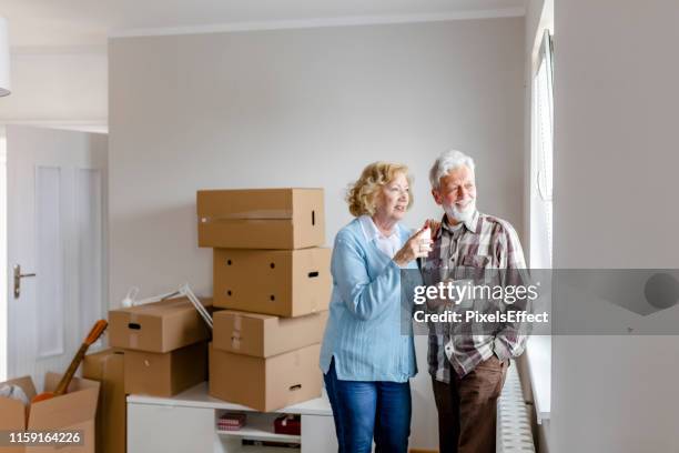 pareja senior en el día de mudanza mirando a través de la ventana - estrenar casa fotografías e imágenes de stock