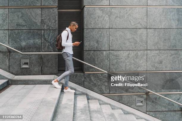 young adult businessman in the city going down the stairs with smart phone - man walking in city stock pictures, royalty-free photos & images