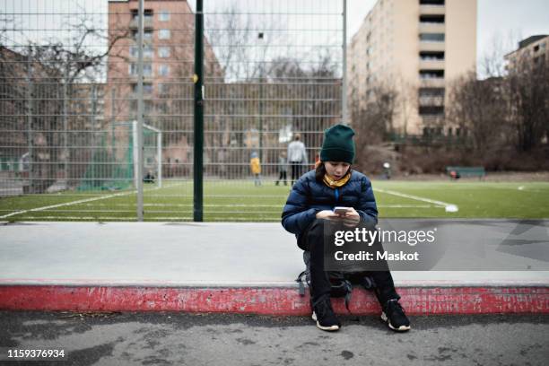 full length of boy wearing warm clothing while using mobile phone against soccer field in city - boy sitting stock pictures, royalty-free photos & images