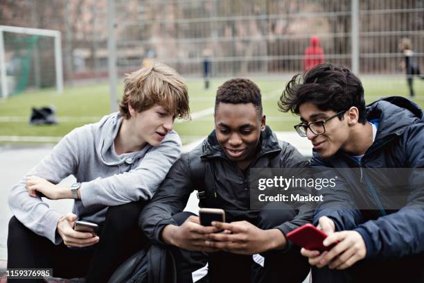 smiling friends looking at teenage boy's phone while sitting against soccer field - mobile phones group stockfoto's en -beelden