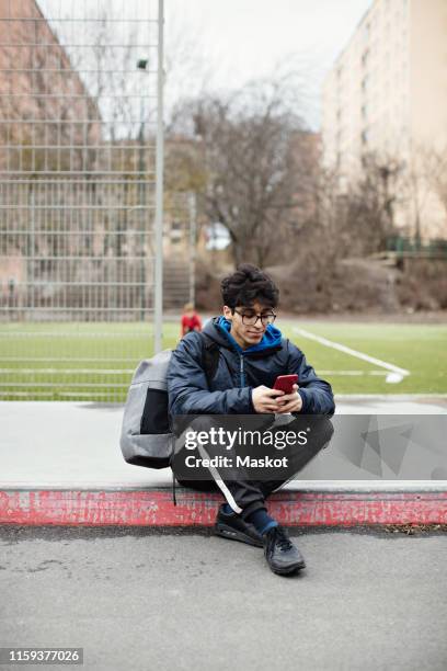 full length of young man using mobile phone while sitting on sidewalk against soccer field during winter - guy with phone full image ストックフォトと画像